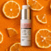 Sérum facial de Vitamina C posicionado sobre fundo laranja vibrante decorado com fatias de laranja frescas, representando ação antioxidante e luminosidade para a pele.